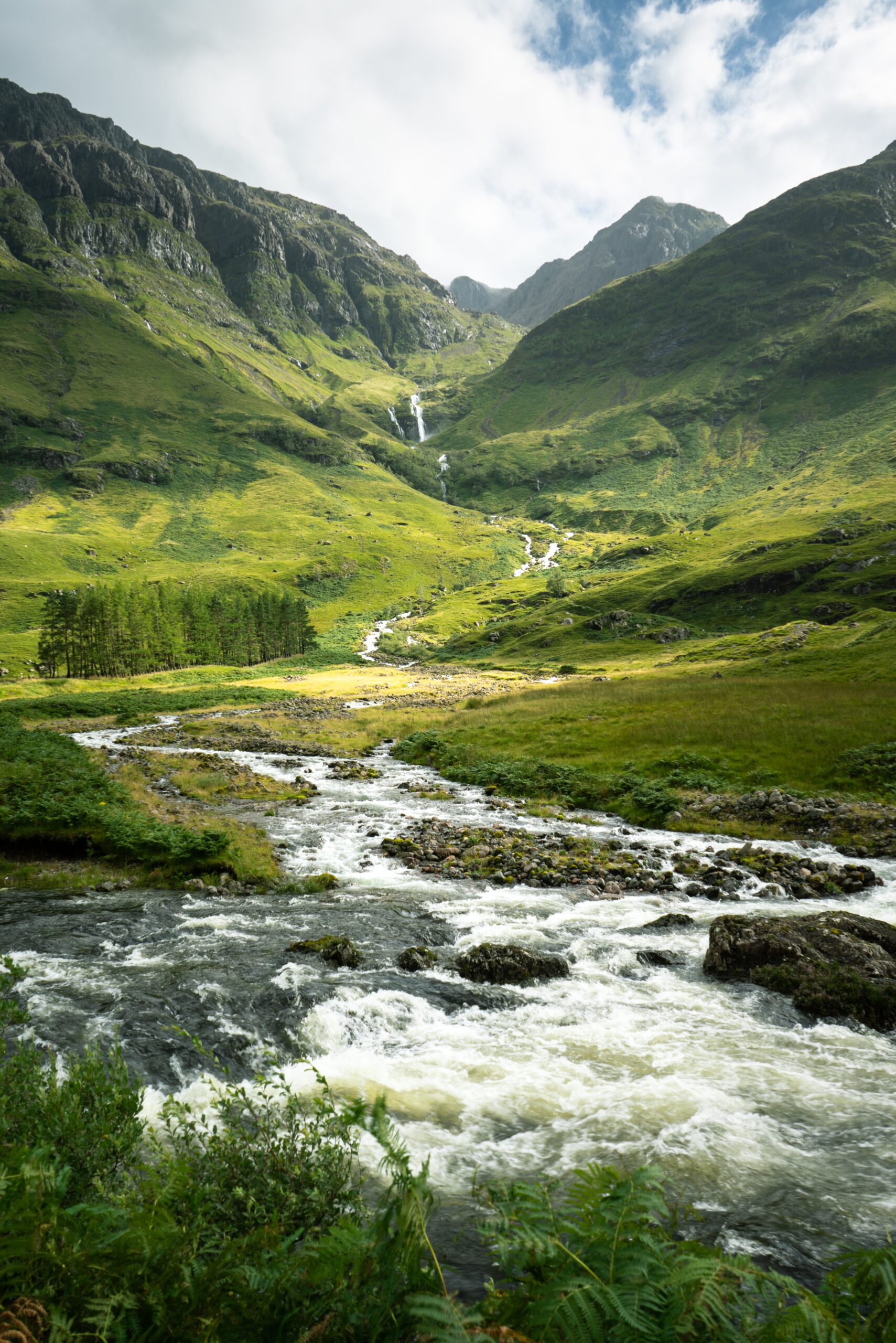 A vertical shot of a river surrounded by the mountains and meadows in Scotland